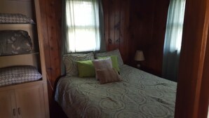 Ein Schlafzimmer mit Bett, Kissen, einem Nachttisch und einer Lampe. Ein Fenster mit Vorhängen und ein Regal mit gemusterten Kissen.
