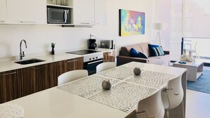 Een moderne keuken met witte kastjes, een magnetron en een koffiezetapparaat. Er is een eethoek met een tafel en stoelen, en een woonkamer met een bank en een kleurrijk abstract schilderij.
