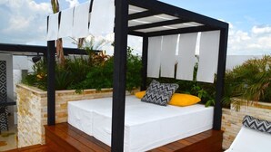 Een overdekt terras met houten vloer, witte zachte zitplaatsen en een pergola.