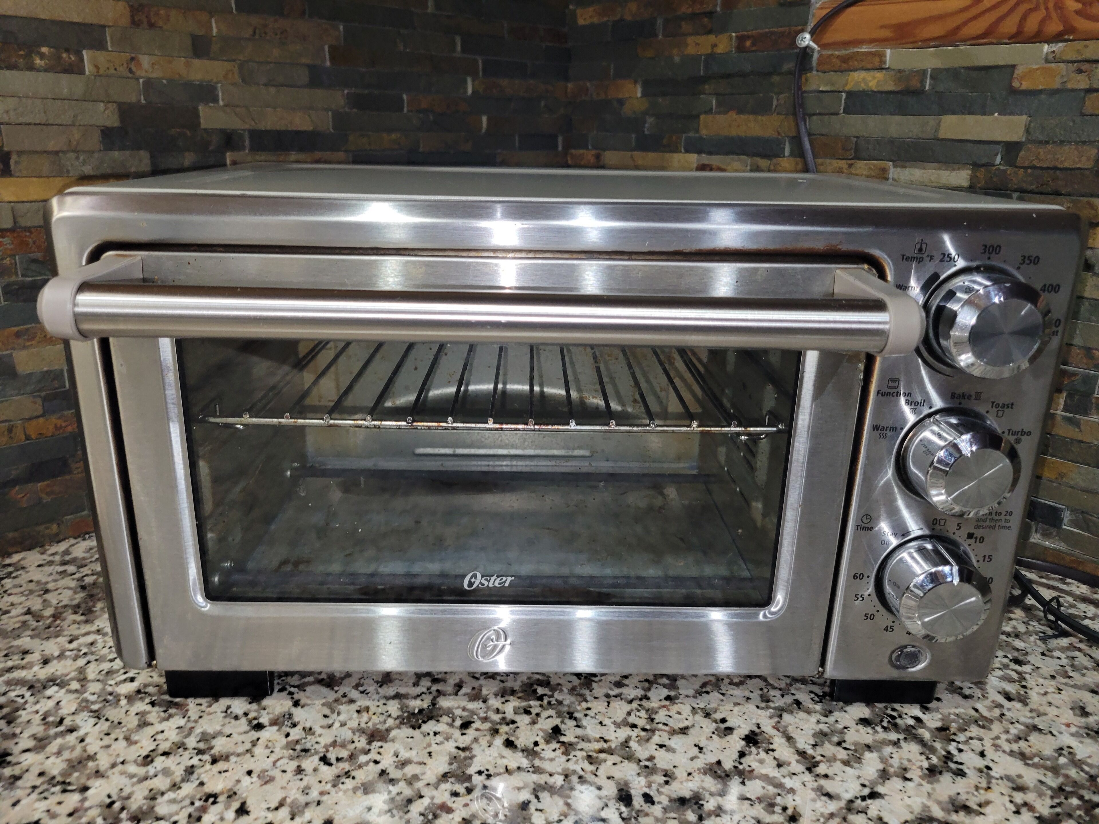Un four grille-pain Oster en acier inoxydable doté d’un cadran de contrôle numérique de la température, d’une minuterie et d’une molette de réglage pour les fonctions telles que le grillage, la cuisson et le toast.
