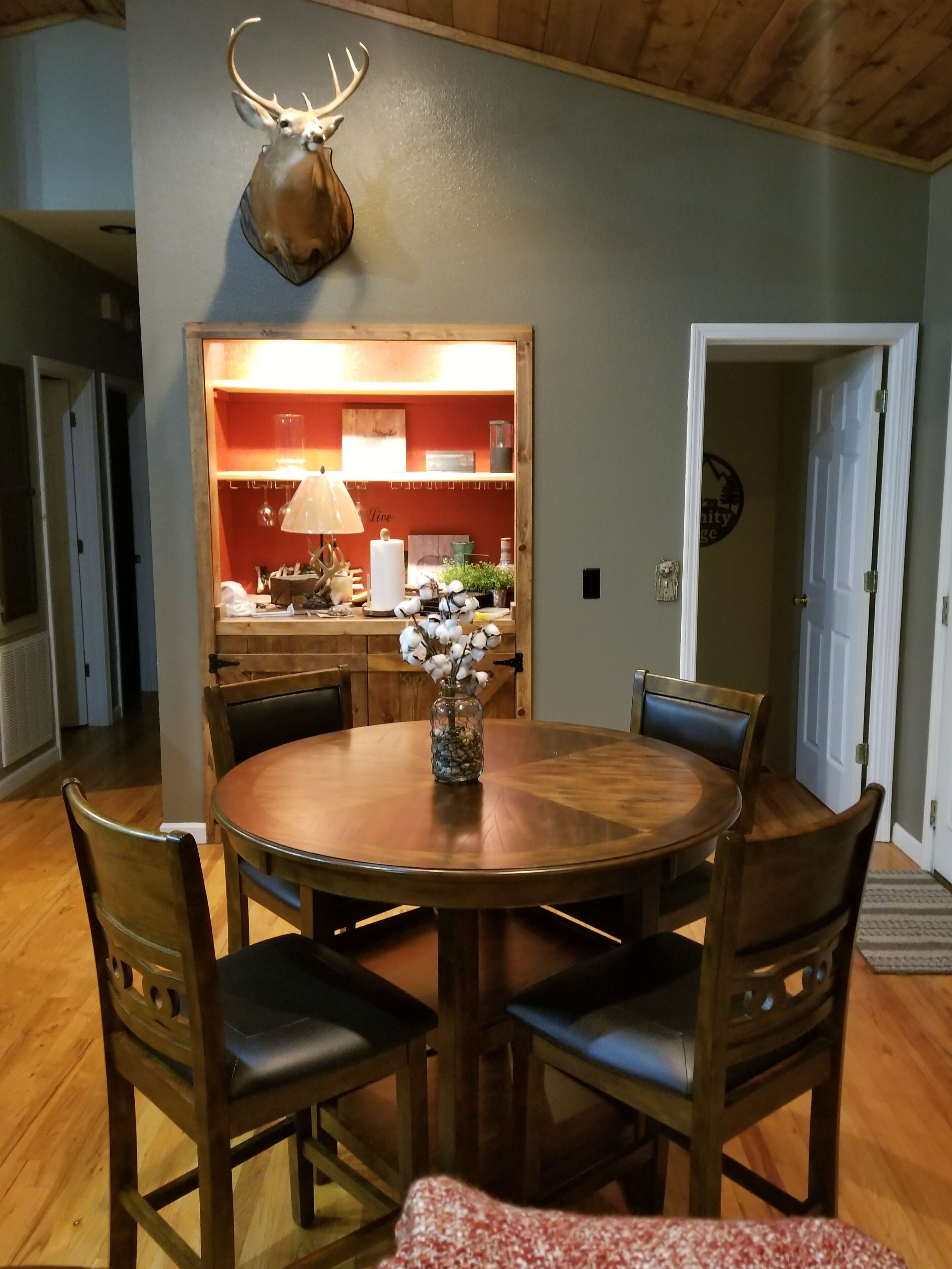 A dining room with a round wooden table, six chairs, and a vase with flowers. A deer head is mounted on the wall above the table.