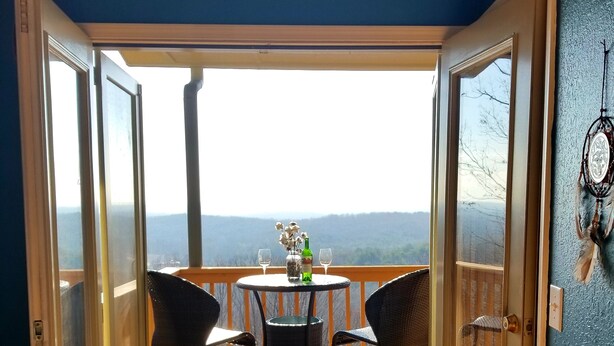 Une porte ouverte donnant sur un balcon avec une table et des chaises, un vase rempli de fleurs et une vue sur le paysage.