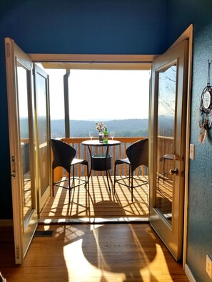 An open door leading to a balcony with a table and chairs, a vase with flowers, and a view of the landscape.