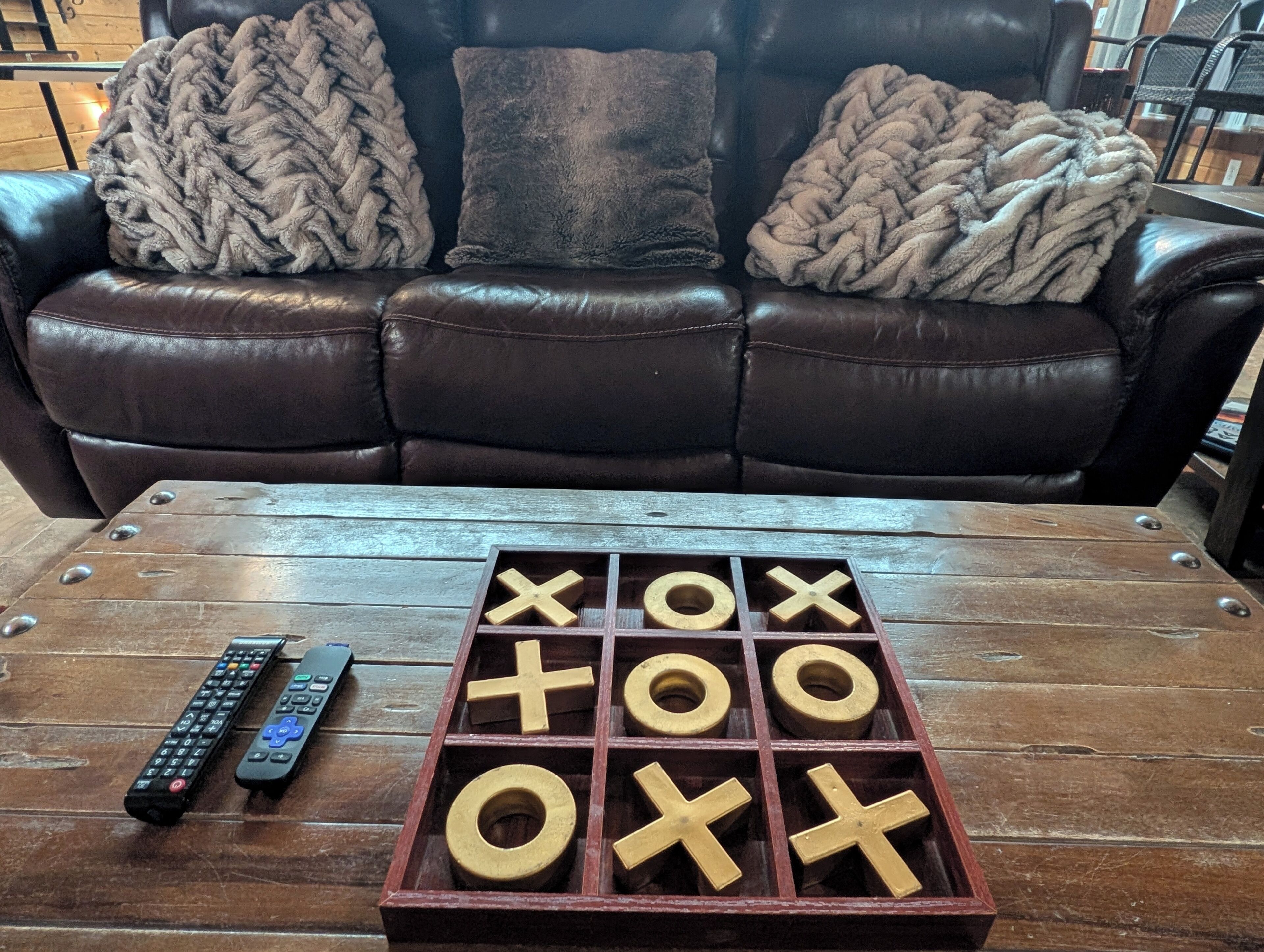 A brown leather couch with three blankets, a wooden coffee table with a tic-tac-toe game and two remote controls.