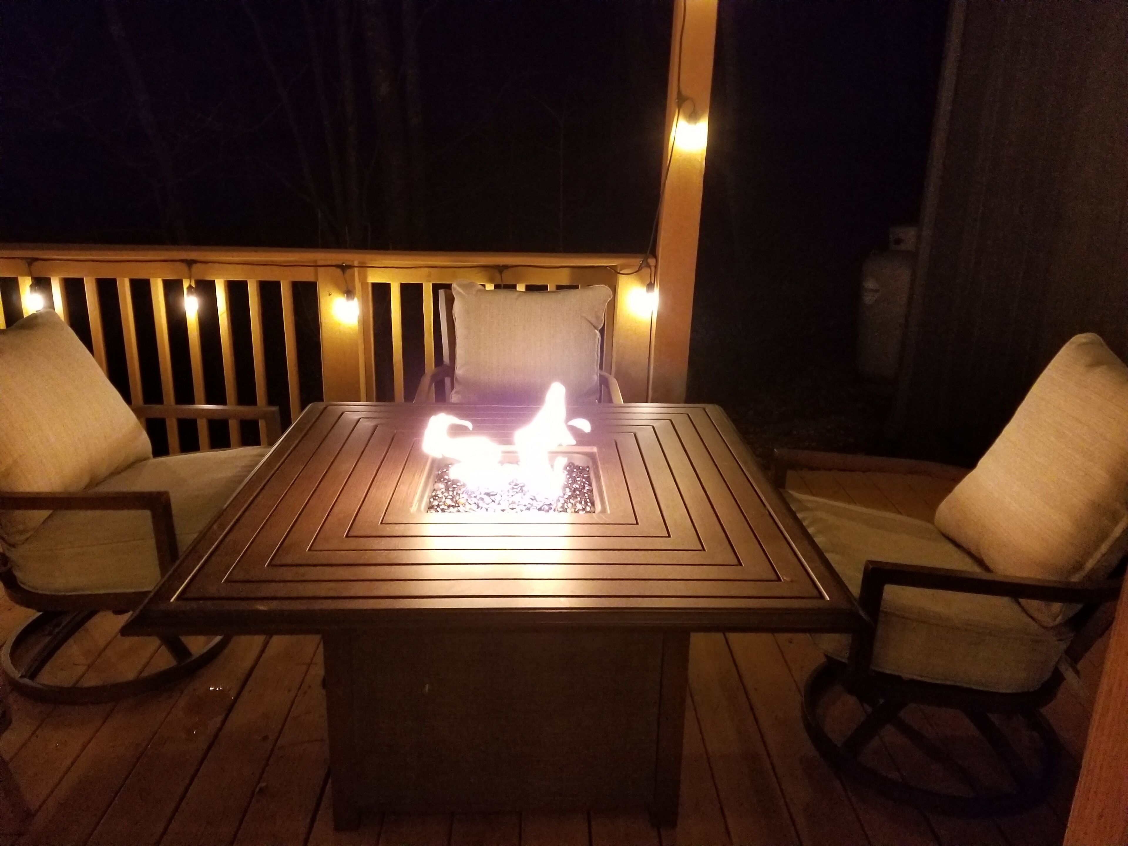 Eine Terrasse im Freien mit einem rechteckigen Tisch und zwei Stühlen, einer Feuerstelle in der Mitte und einem Holzgeländer mit Beleuchtung.