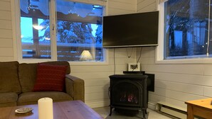 Ett mysigt vardagsrum med ett träbord, ett vitt ljus, en brun soffa med en röd kudde, en svart tv på väggen och en vedkamin.