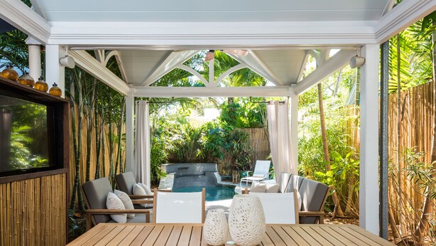Un espacio al aire libre techado con una mesa y sillas de madera, piscina y abundante vegetación.