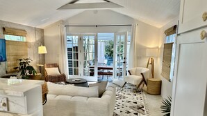 Amplia sala de estar con sofá, mesa de centro y mesa de comedor. Se observa un gran ventanal con cortinas, una lámpara y una planta.