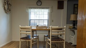 Un comedor con una mesa de madera y cuatro sillas, una ventana con persianas, un reloj de pared y una lámpara en una mesita.