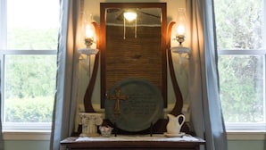 Un dormitorio con un aparador de madera, un espejo y dos apliques en la pared. Se ve una ventana con cortinas y vistas al exterior con vegetación.