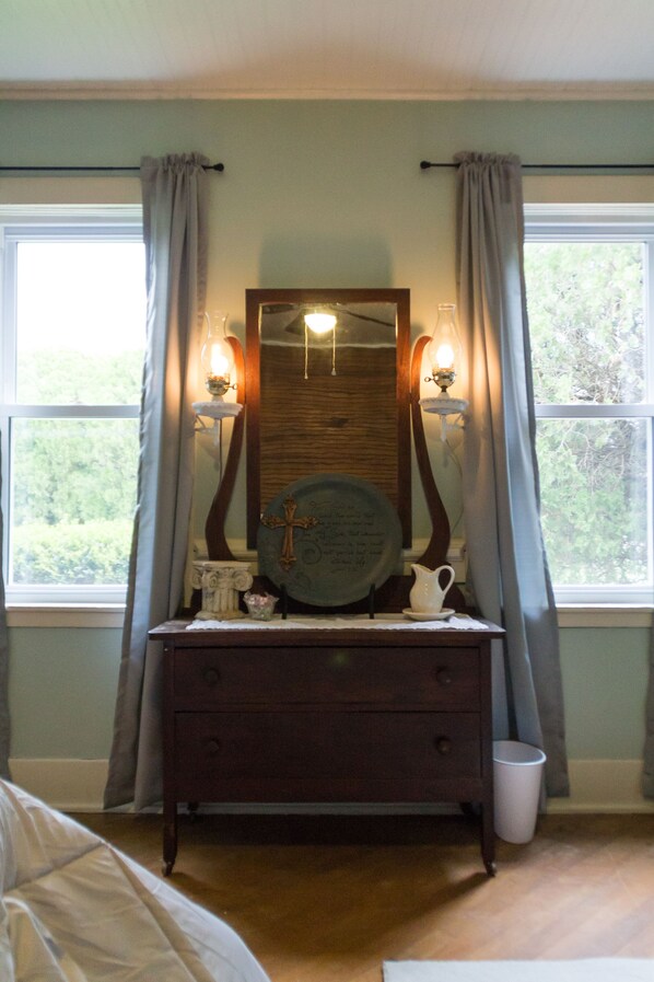 A bedroom with a wooden dresser, a mirror, and two wall sconces. There is a window with curtains and a view of greenery outside.
