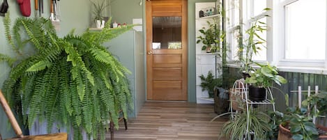 A wooden-floored hallway with a wooden door, potted plants, and a variety of hanging kitchen utensils.