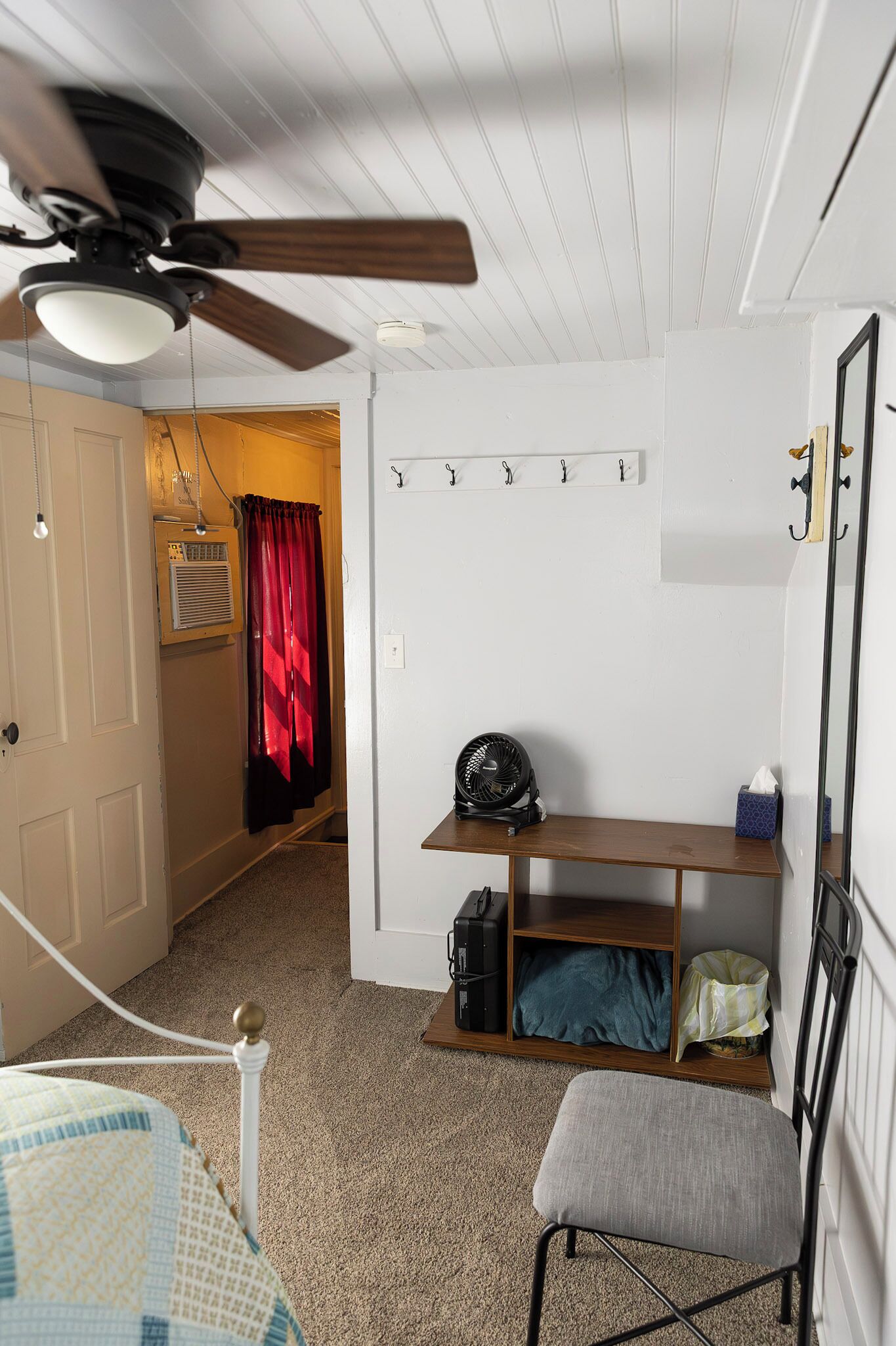 Une pièce avec un ventilateur de plafond, un lit, un banc avec une valise, un radiateur et un miroir au mur.