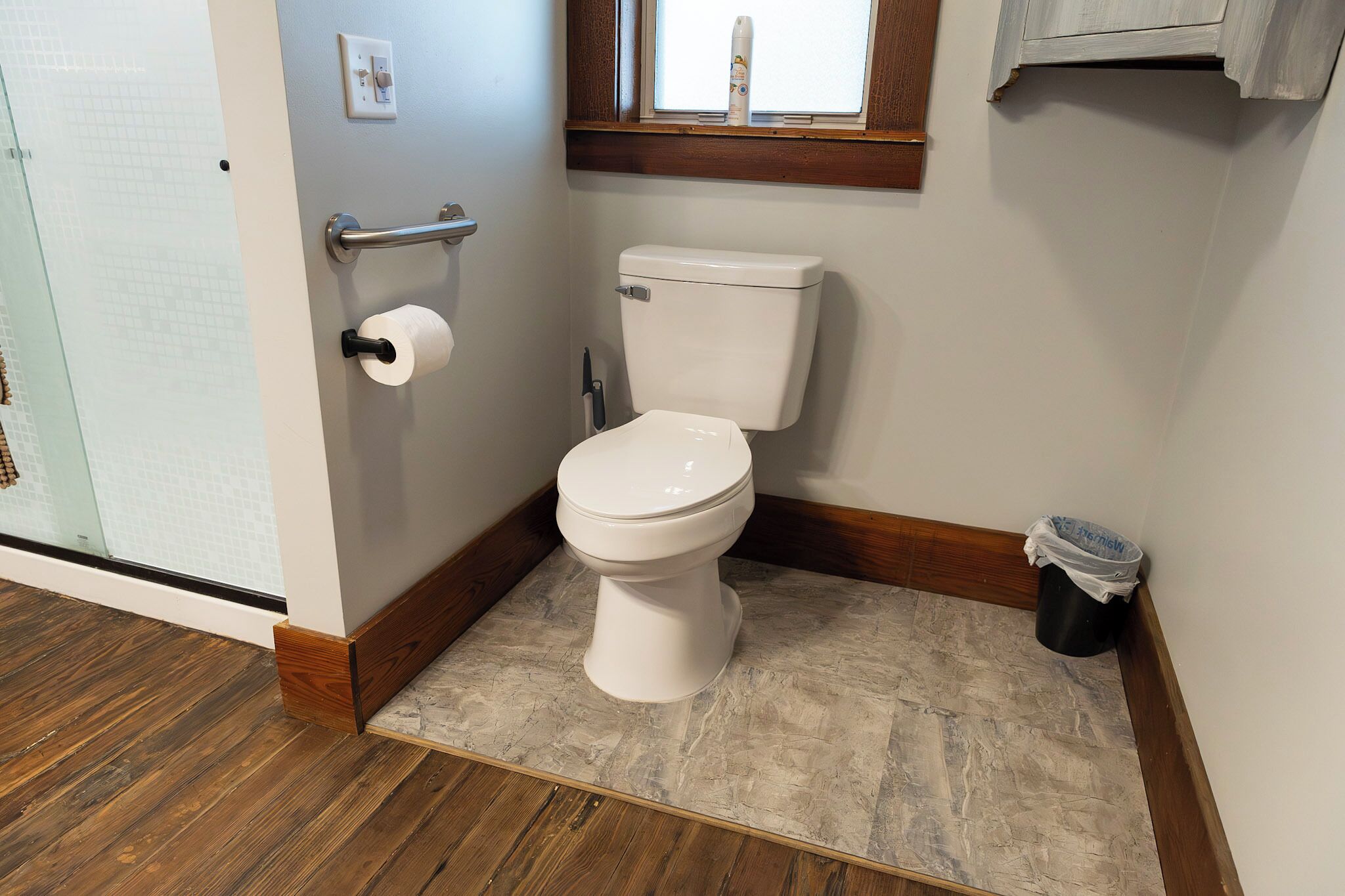 A bathroom with a toilet, a roll of toilet paper, a window, and a trash can.