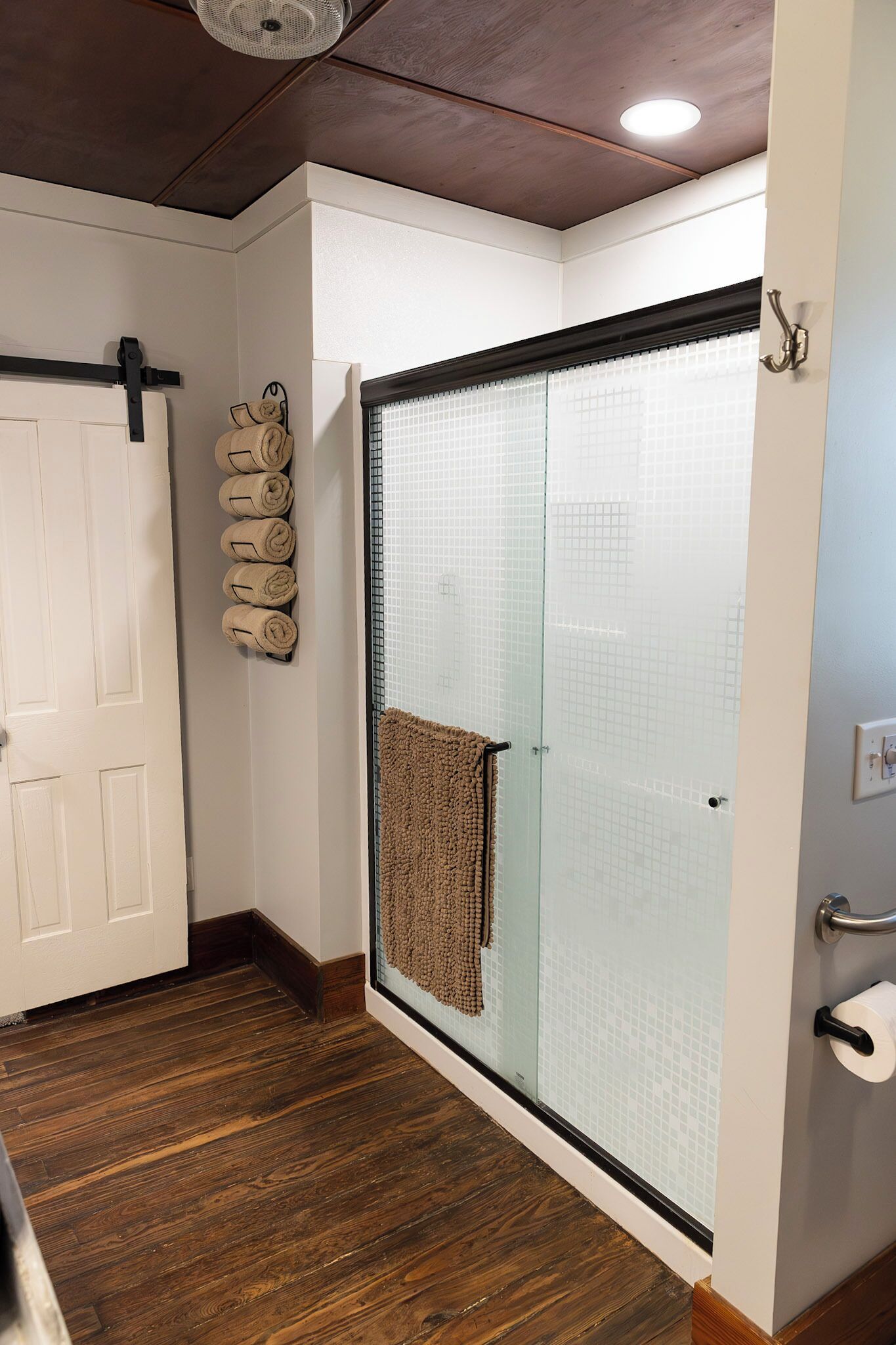 Une salle de bain avec une porte de douche en verre, un porte-serviettes avec des serviettes, un plancher en bois et une porte blanche.