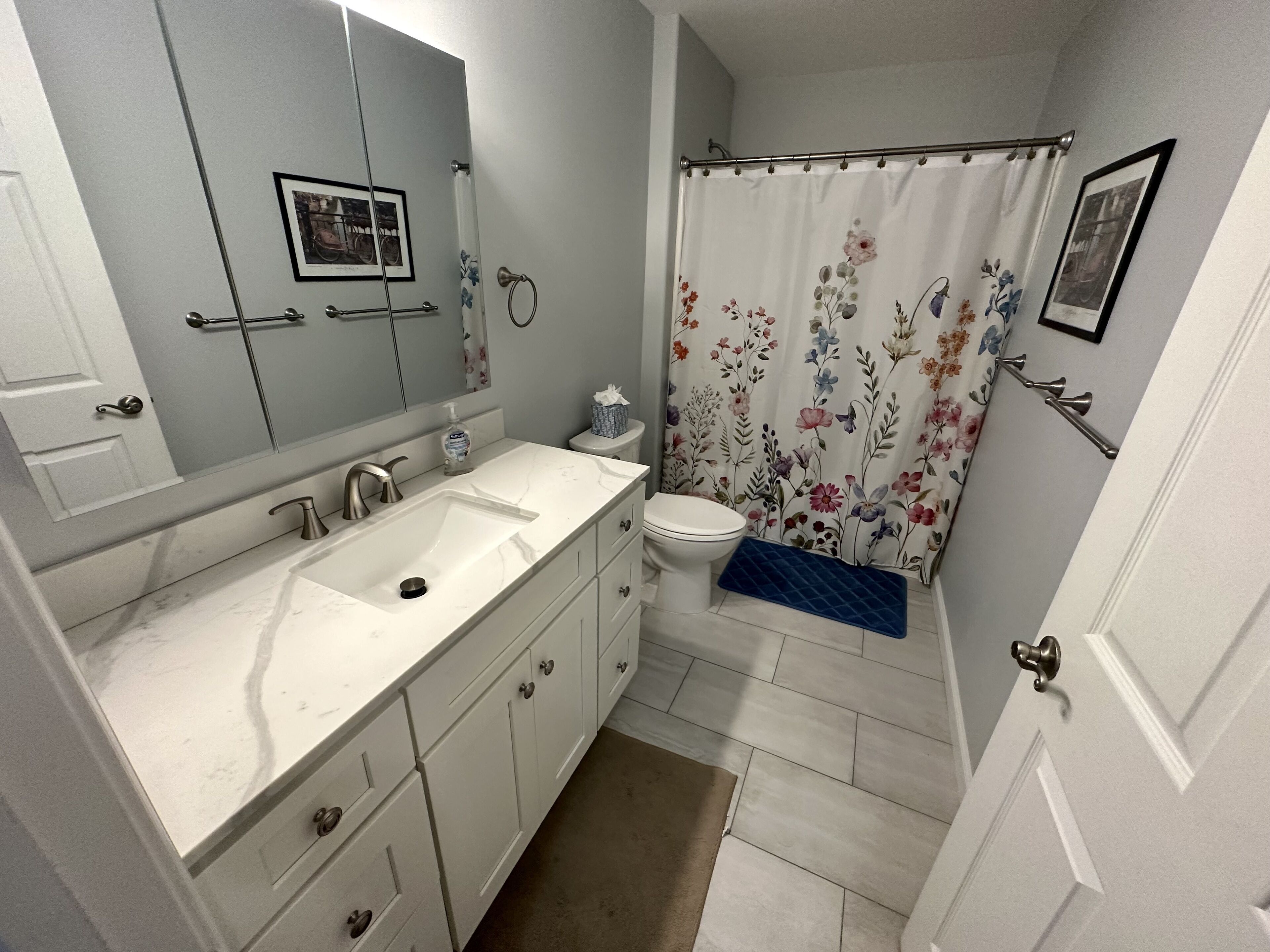 A bathroom with a white sink, mirror, and toilet. There is a shower curtain with floral patterns and a blue bath mat.