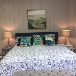 Ein Schlafzimmer mit einem gemusterten Bettbezug, einem Kopfteil mit drei Zierkissen und einem Bild über dem Bett.