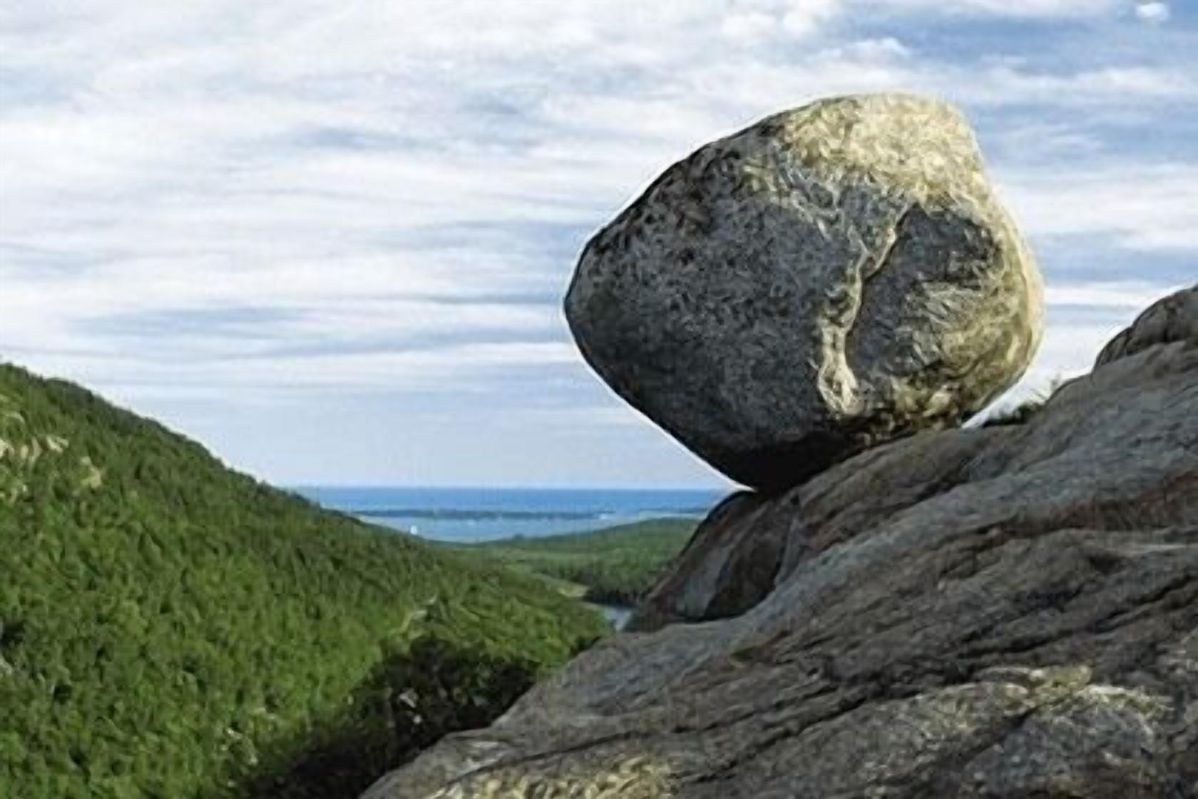 A large rock balanced on another rock with a view of a green valley and a body of water in the distance.