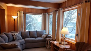 A cozy living room with a sectional sofa, a wooden coffee table, and a chair. Large windows with curtains provide a view of trees outside. - Lake Superior Home - Perfect for a family vacation any time of the year! (Calumet)