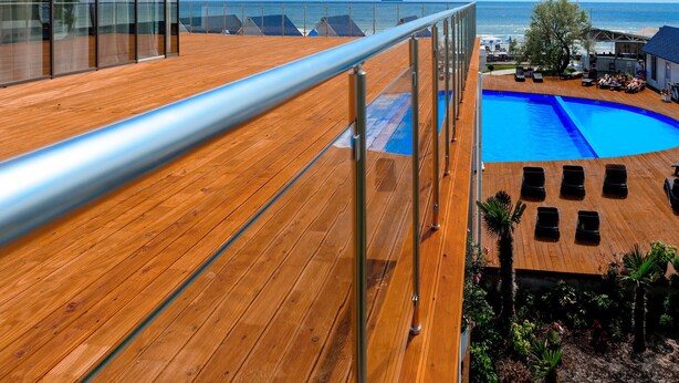 A wooden deck with a glass railing overlooking a pool and the sea.