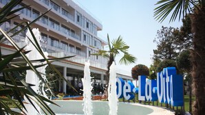 A fountain with water jets in a circular pool, surrounded by palm trees and a building with a sign that reads "De La Violette".