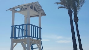Een uitkijktoren voor strandwachten met een ladder op een zandstrand, palmbomen en een helderblauwe lucht.