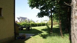 Un jardin à l’arrière d’une maison, avec une terrasse dallée, une table en bois et des plantes en pot.