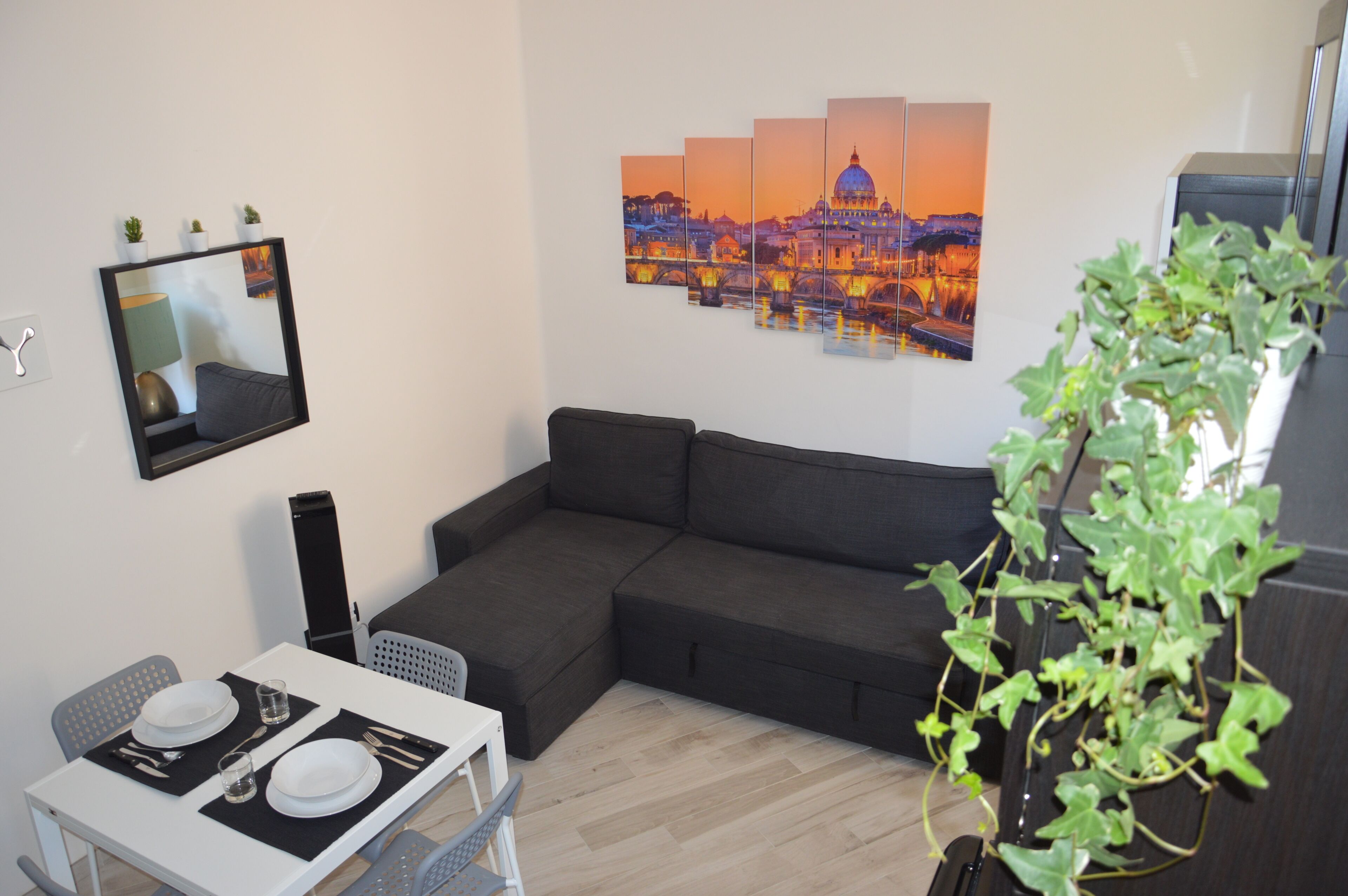 Un salon moderne avec un canapé d’angle, une table à manger pour deux personnes, un téléviseur à écran plat fixé au mur et un grand tableau représentant un paysage urbain, placé au-dessus du canapé.