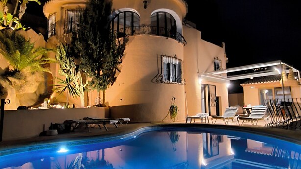 A nighttime view of a hotel with a swimming pool, a multi-story building with balconies, and outdoor seating.