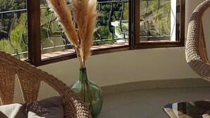 A room with a large window offering a view of a mountainous landscape and the sea. There is a wicker chair and a glass table with a vase of pampas grass.