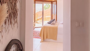 A hallway leading to a bedroom with a bed, a hanging woven light fixture, and a decorative wall piece.