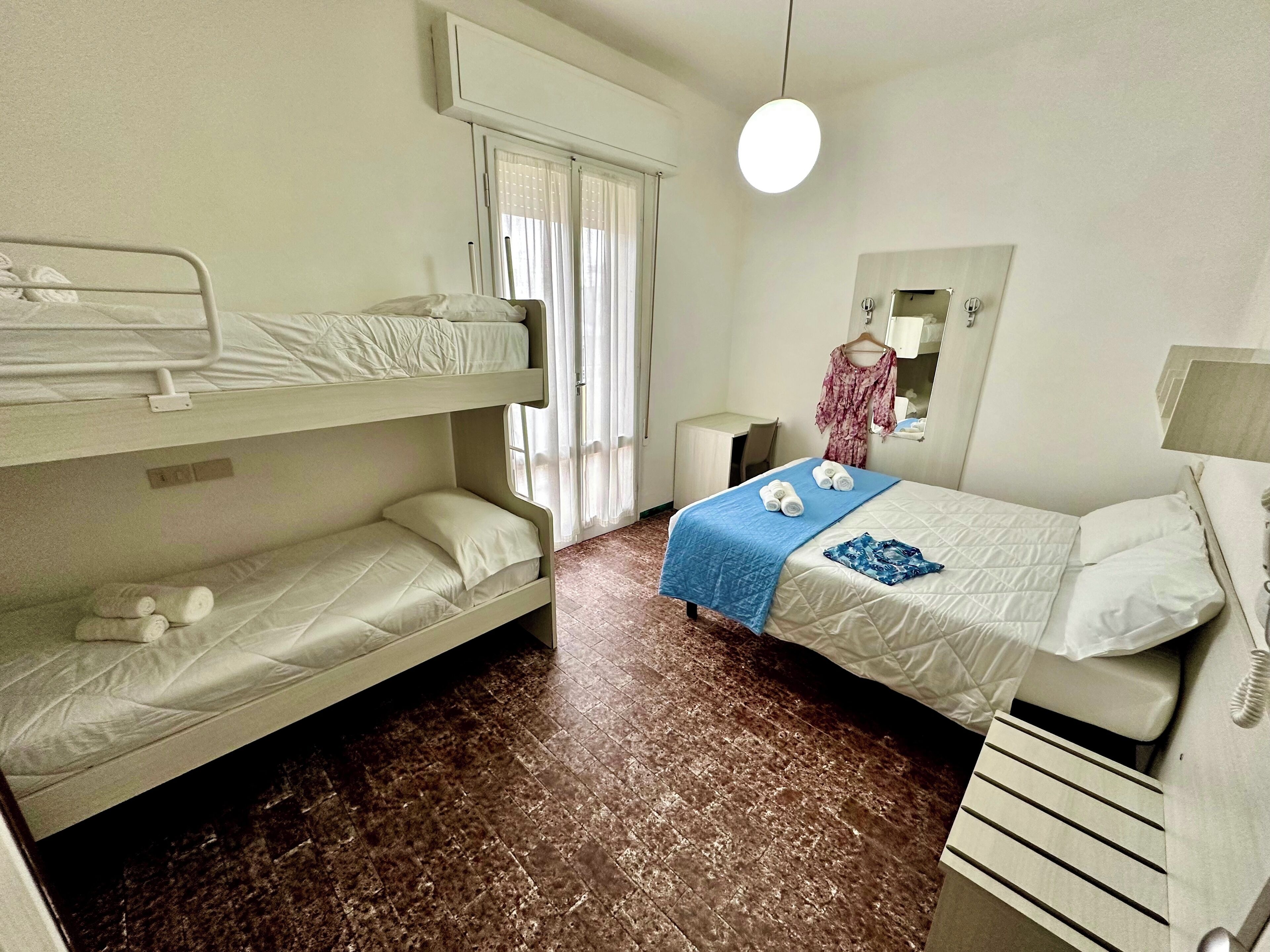 Ein Zimmer mit Etagenbetten, einem Schreibtisch, einem Stuhl, einem hängenden Kleid, einer Lampe und einem Telefon.