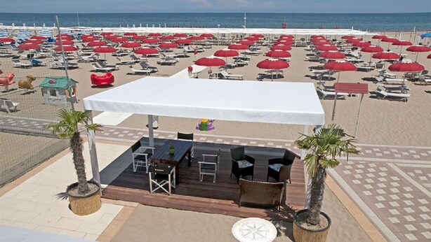 A beachside restaurant with outdoor seating, a white canopy, and palm trees.