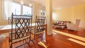 A dining area with a wooden table and chairs, a sofa, and a striped armchair in a bright room with large windows. - Spacious and bright designer apartment next to Playa Larga (Tarragona)