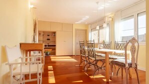 A spacious dining area with a wooden table and chairs, a built-in bookshelf, and large windows allowing natural light. - Spacious and bright designer apartment next to Playa Larga (Tarragona)