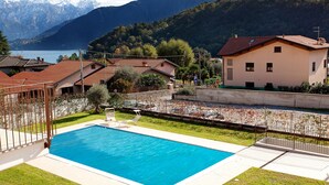 A swimming pool with a wooden deck, surrounded by a metal fence, set against a backdrop of mountains and a lake. - Lenno Spese II –Village apartment In Lenno with pool by Italian Apartments. (Lenno)