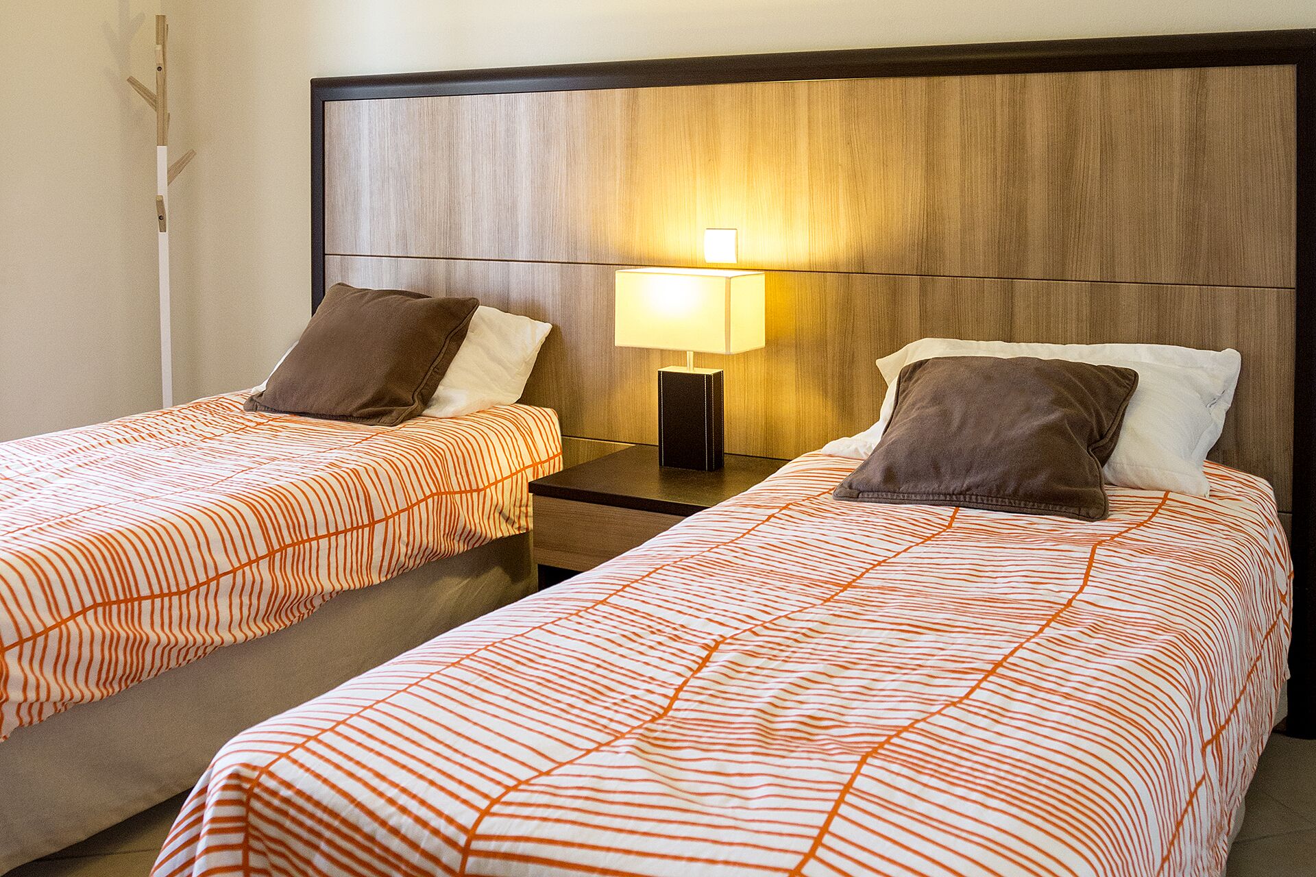 Twee enkele bedden met oranje streepjes bekleed, een houten hoofdbord, een nachtkastje met een lamp, en een wandlamp.