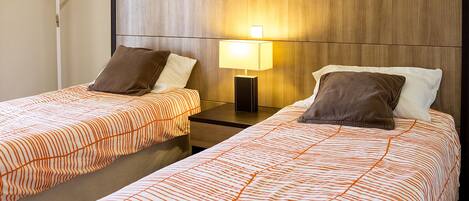 Twee enkele bedden met oranje streepjes bekleed, een houten hoofdbord, een nachtkastje met een lamp, en een wandlamp.