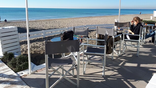 An outdoor seating area with tables and chairs, overlooking a beach and the sea.