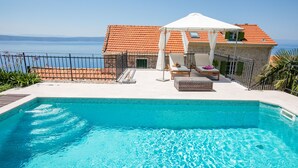 Une piscine avec vue sur la mer, un espace de détente couvert avec des chaises, et un bâtiment en pierre avec un toit en tuiles.