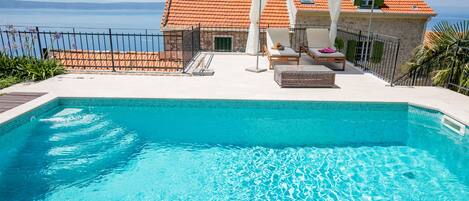 A swimming pool with a view of the sea, a covered seating area with chairs, and a stone building with a tiled roof.
