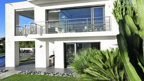 Una casa moderna de dos pisos con balcón, piscina y jardín con cactus y palmeras.