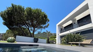 Un área de piscina moderna con agua cristalina, rodeada por un edificio blanco con balcón y un gran árbol al fondo.
