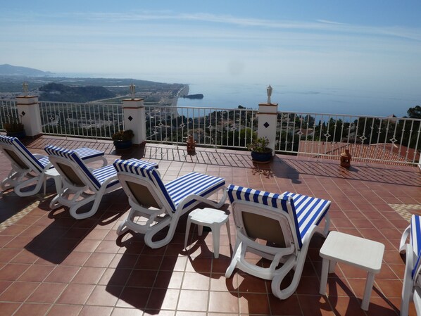Una terraza con sillones blancos, mesas, plantas en macetas y vistas al mar y al paisaje lejano.