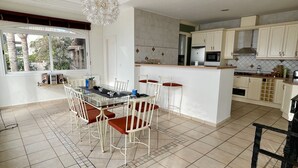 Une cuisine moderne avec des meubles blancs, un coin repas doté d’une table en verre et de chaises blanches, et une vue sur les palmiers par la fenêtre.