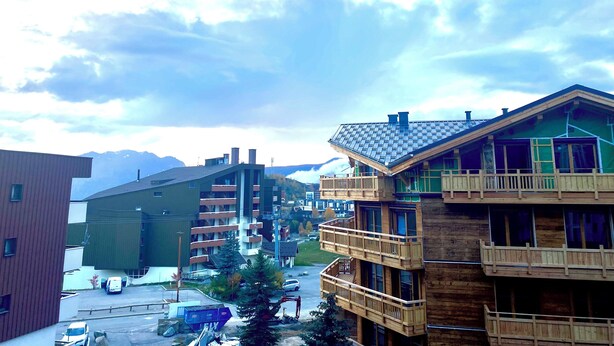 A multi-story building with balconies, a parking area, and a mountain backdrop.