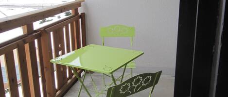 A balcony with a green metal table and chair, a wooden railing, and a view of buildings in the background.