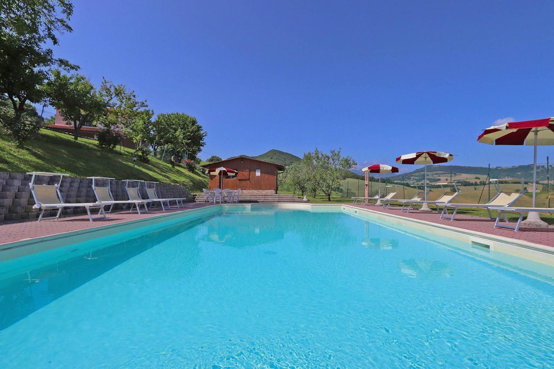 Una piscina con sillones de playa y sombrillas, un edificio de madera y un paisaje montañoso al fondo.