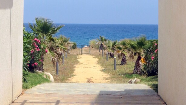 Un sendero conduce a una playa con vista despejada al océano, rodeada de vegetación y una estructura de pérgola.