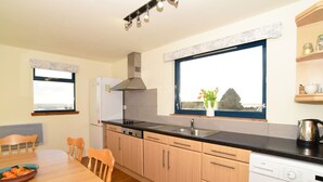 A compact kitchen with wooden cabinets, a black countertop, a built-in dishwasher, and a window with a view. - Cottage With Secure Garden, Wood Burning Stove And Sea Views (St Cyrus)
